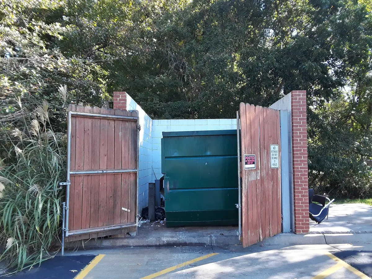A green dumpster inside a block-wall enclosure with its wooden gates open, at the edge of a parking lot