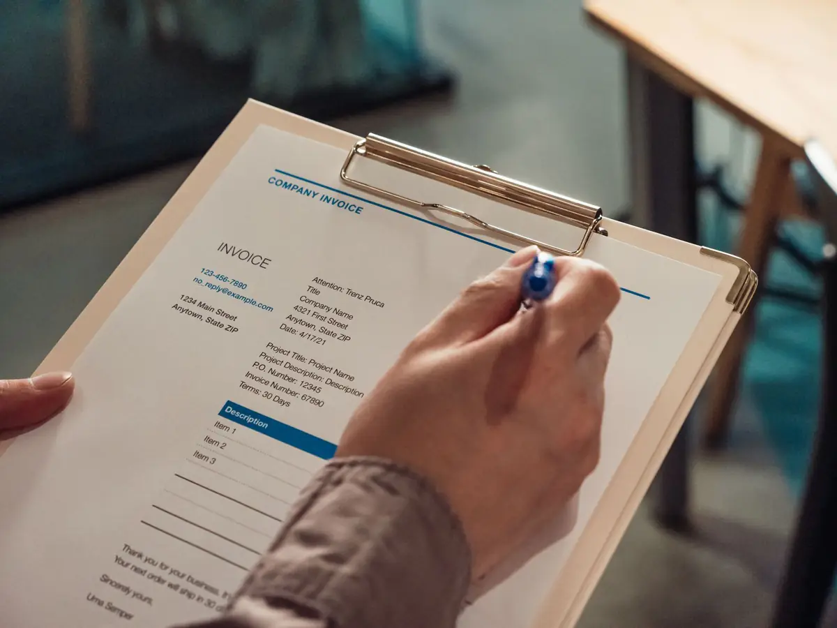 A hand filling in an invoice on a clipboard with a pen