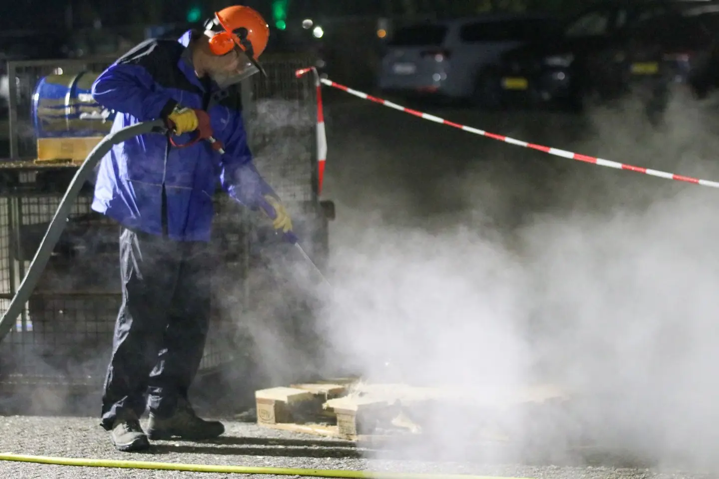 A worker in a helmet, face visor and ear protection pressure washing at night as steam rises from the spray