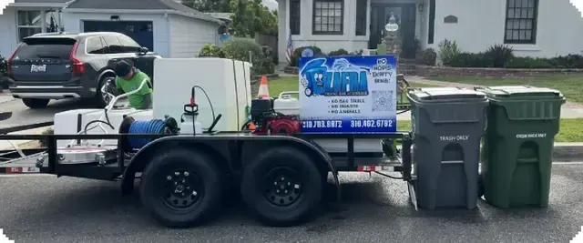 An open utility trailer with white water tanks, a red pressure washer and a hose reel, parked at the curb beside two carts